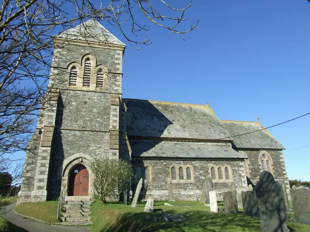 The exterior of St John the Evangelist church in Delabole, with its grey slate tower and the churchyard under a clear blue sky