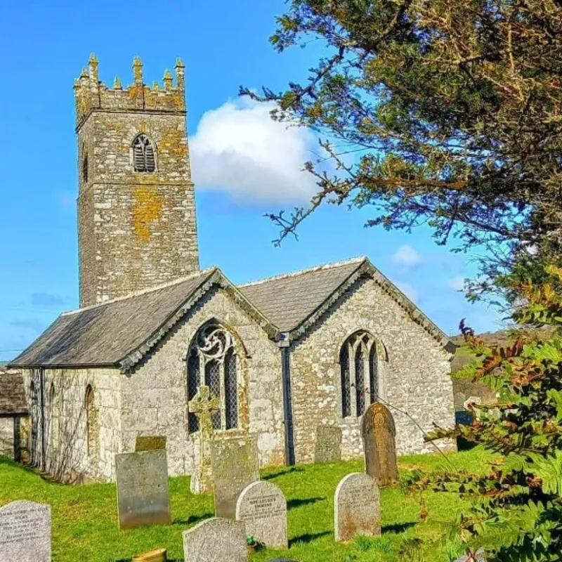St Adwena’s church and tower at Advent, seen from the green churchyard with gravestones under a bright blue sky